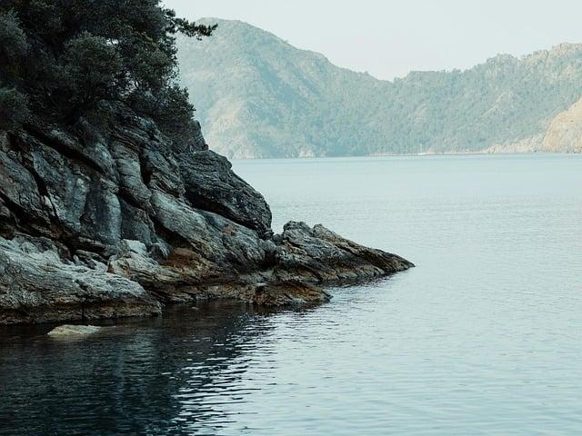 A peaceful lakeside scene with rocky cliffs covered in greenery and calm blue water reflecting the distant misty mountains.